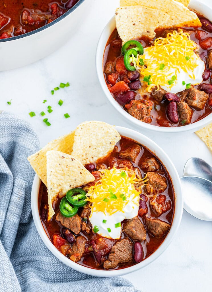 Two bowls of steak chili topped with tortilla chips, sour cream, and cheddar cheese.