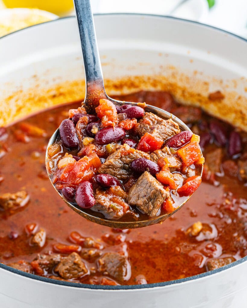A ladle full of steak chili above a pot of it.