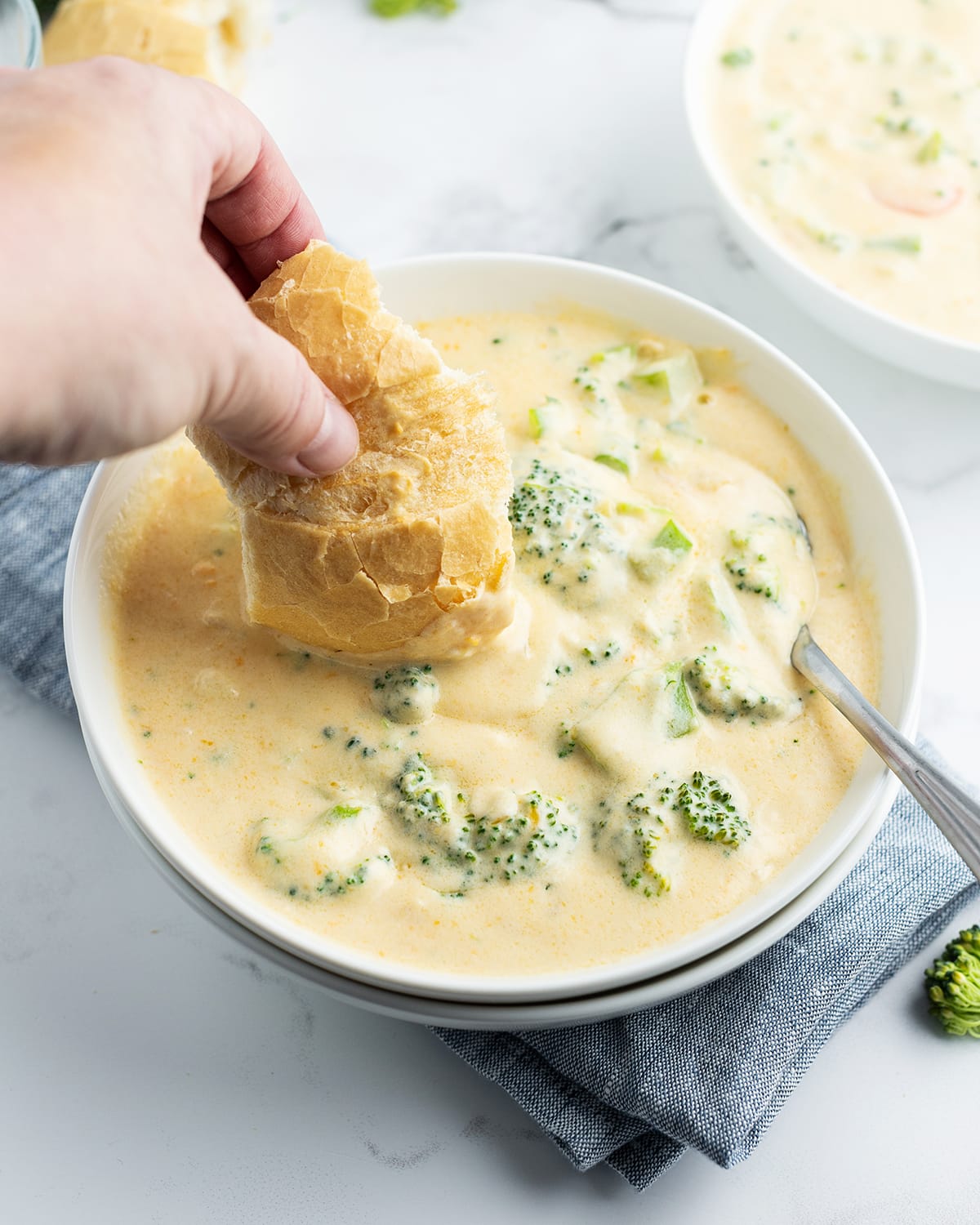 A hand holding a piece of bread dipping into a bowl of broccoli cheese soup.