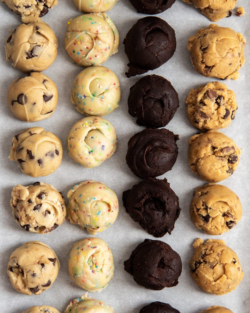 Rows of scoops of different flavors of edible cookie dough on parchment paper. There is chocolate chip cookie dough, sugar cookie dough, chocolate cookie dough, and peanut butter cookie dough.