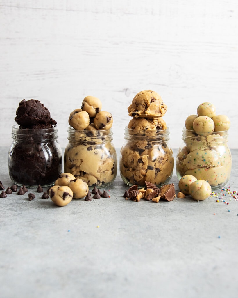 Four jars of cookie dough in a row. There is double chocolate cookie dough, chocolate chip cookie dough, peanut butter cookie dough, and sugar cookie dough.