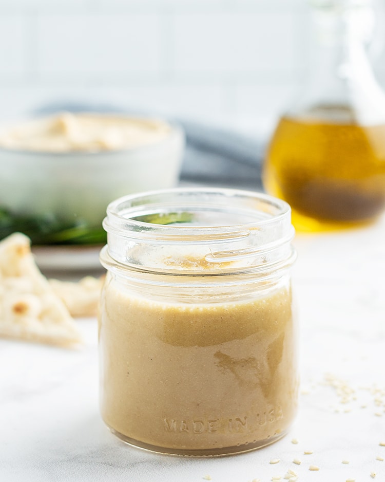 A jar of tahini with a bowl of hummus, and a bottle of oil behind it.