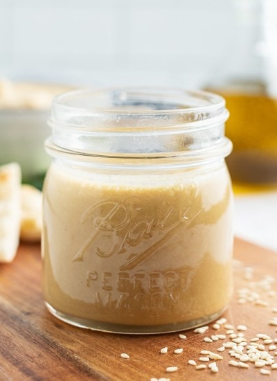 A close up of jar of tahini with a bowl of hummus, and a bottle of oil behind it.