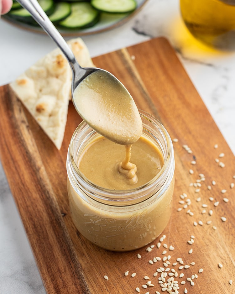 A small jar full of homemade tahini with a spoon above it drizzling the tahini into the jar.