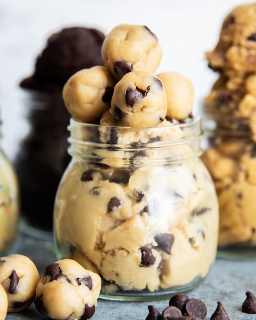 A jar of edible chocolate chip cookie dough topped with small balls of cookie dough, and with small balls in front of the jar too. 
