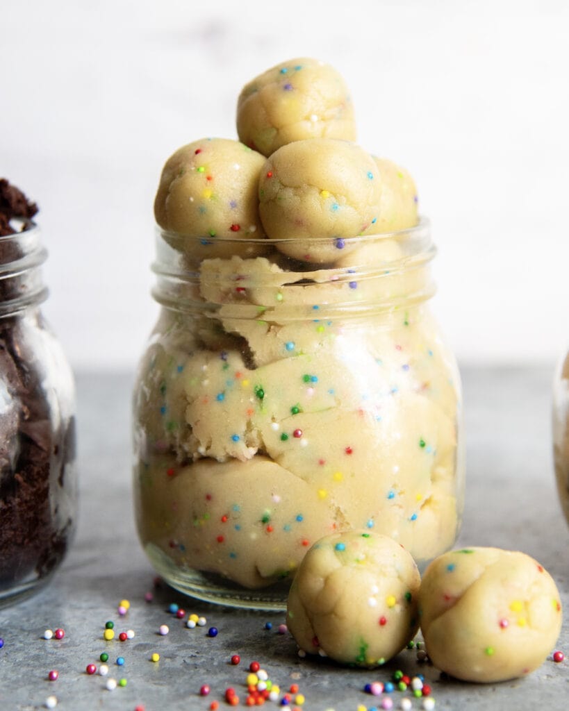 A glass jar full of cookie dough with rainbow sprinkles. The jar is topped with small balls of sugar cookie dough.