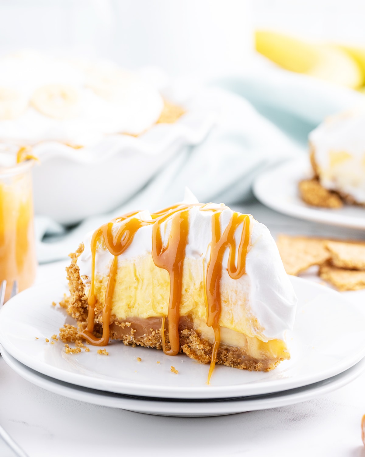 A slice of caramel banana cream pie with a graham cracker crust, caramel, pudding, whipped cream, and drizzled with caramel.