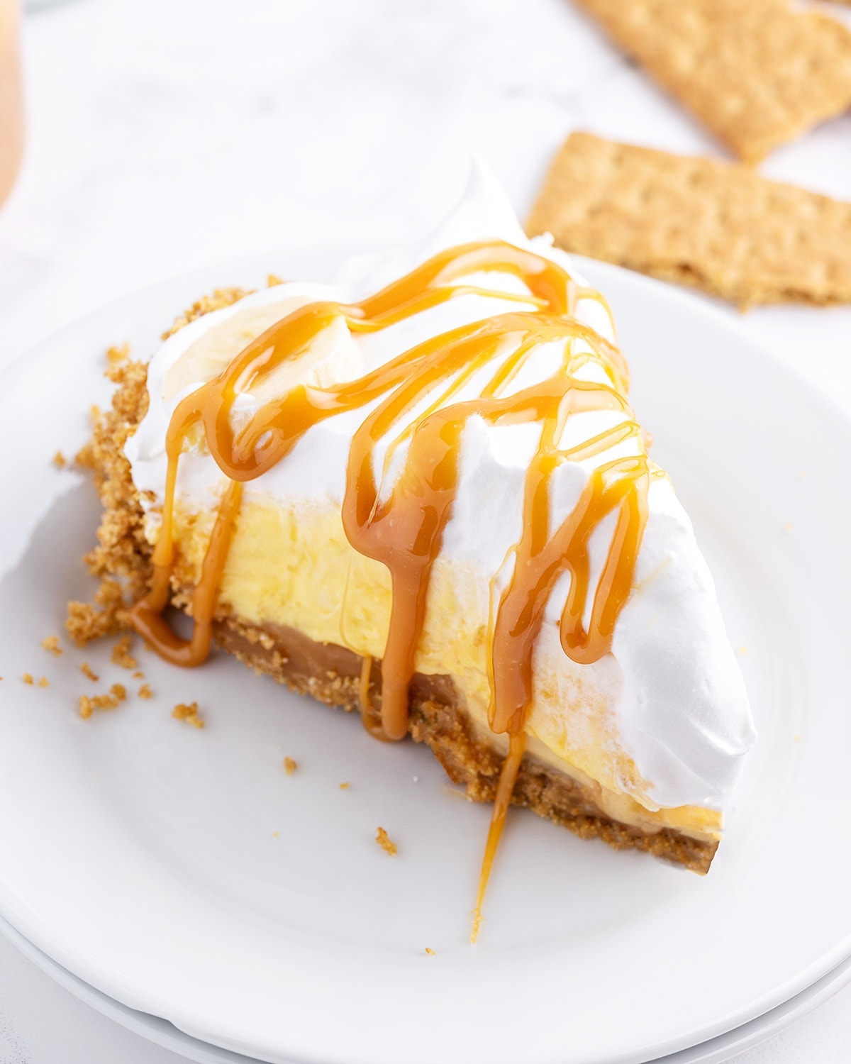 A slice of caramel banana cream pie on a white plate topped with caramel sauce.