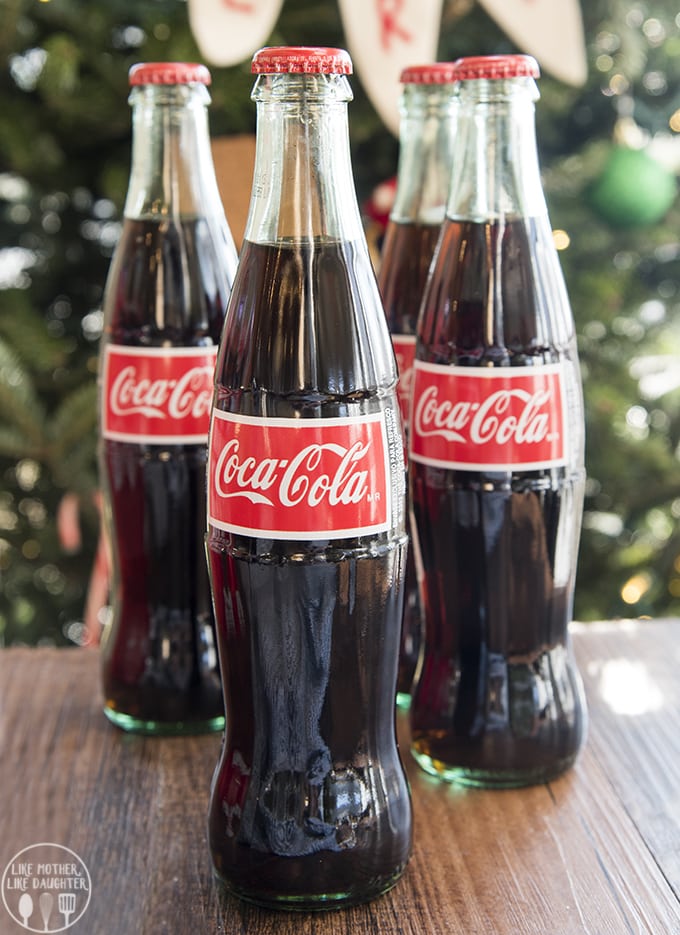 Four glass bottles of coca cola in front of a Christmas tree.