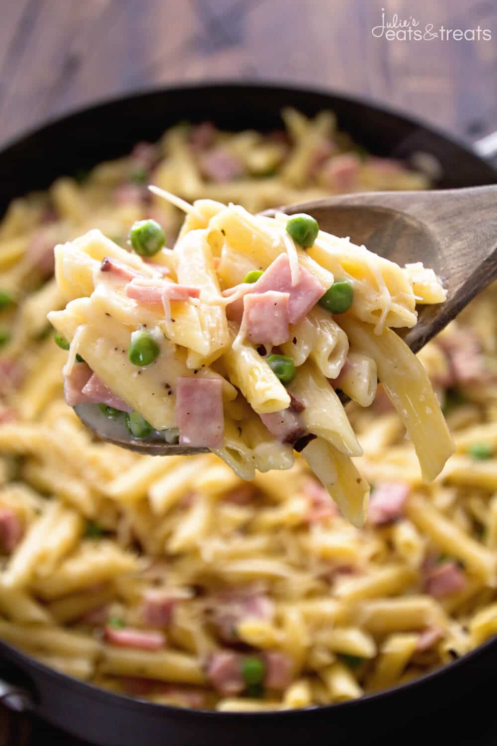 A spoonful of penne, peas, and ham above a skillet. 
