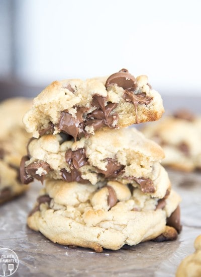 A stack of large chocolate chip cookies, the top cookie is broken in half showing the melted chocolate chips.