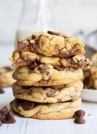 A stack of thick levain style chocolate chip cookies, with one broken in half showing the gooey center on top.