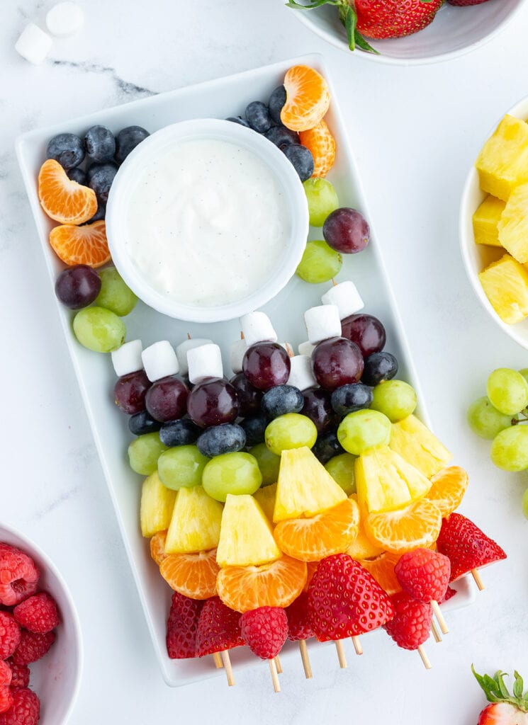 A tray of fruit kabobs decorated in rainbow colors, and a bowl of fruit dip.