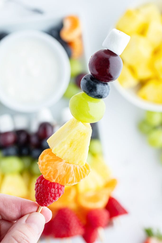A hand holding a fruit skewer with a raspberry, orange, pineapple, grapes, and blueberries.