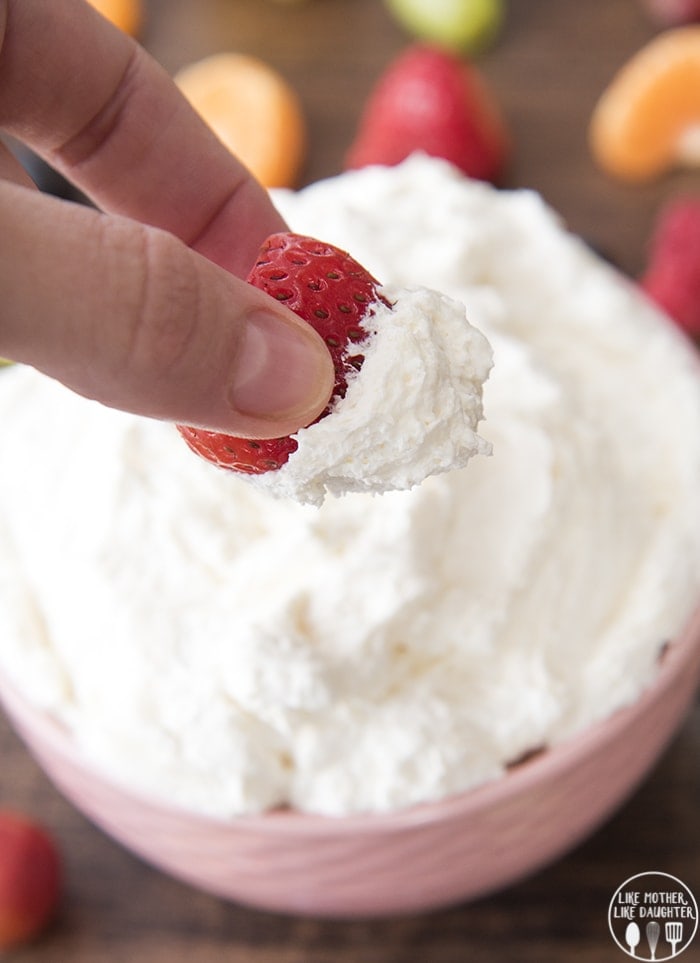 A hand holding a strawberry above a bowl of marshmallow cream cheese fruit dip.