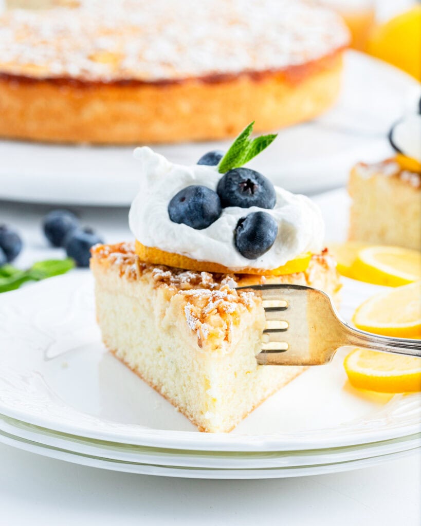 A piece of lemon ricotta cake topped with a dollop of whipped cream, three blueberries, and mint leaves, with a fork digging into the cake.