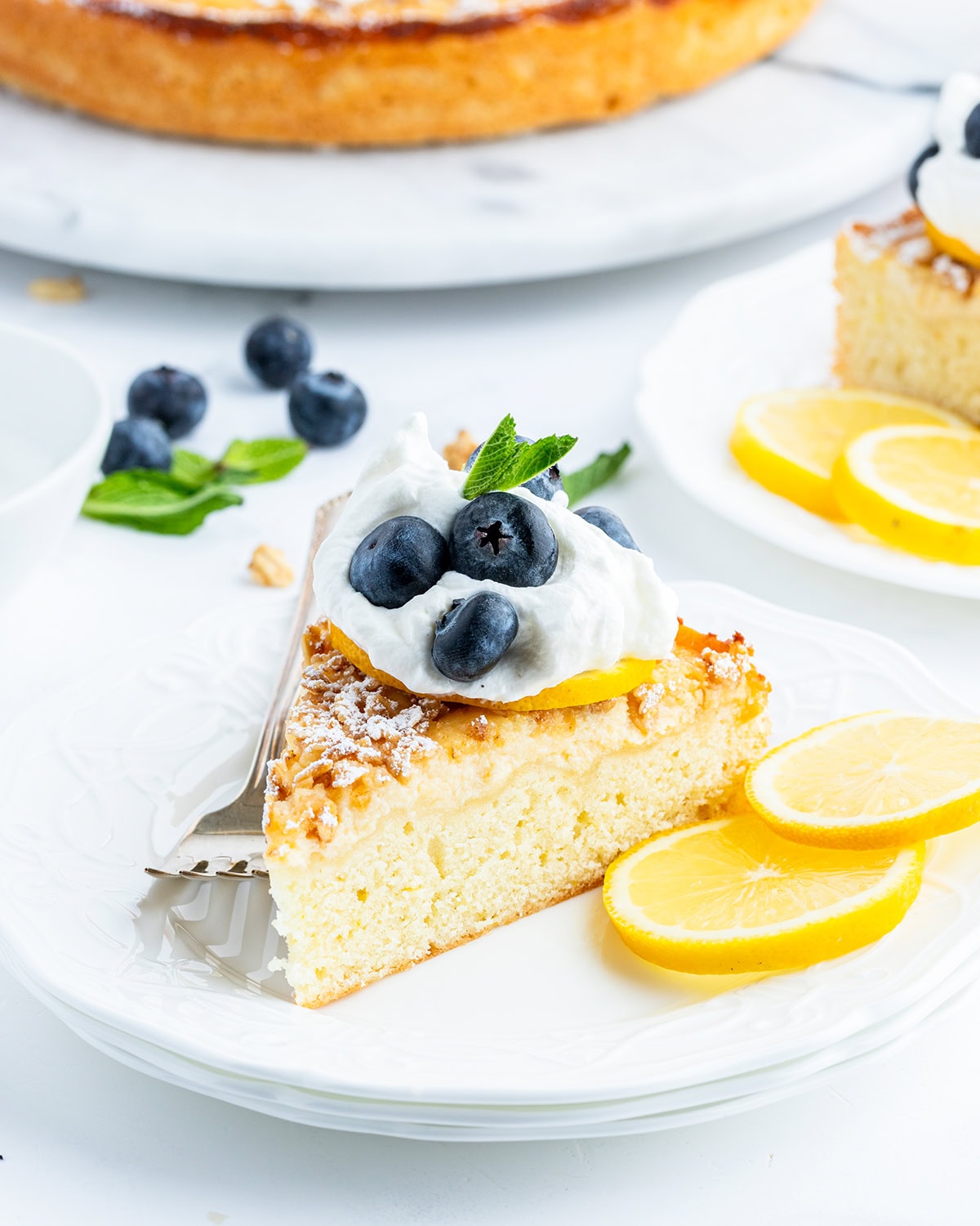 A piece of lemon ricotta cake topped with a dollop of whipped cream, three blueberries, and mint leaves.