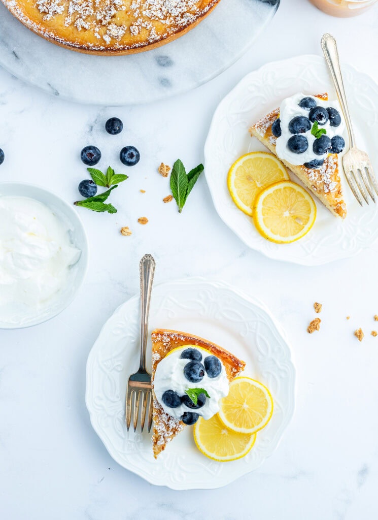 An above view of two plates with slices of ricotta cake, topped with whipped cream and fresh berries.