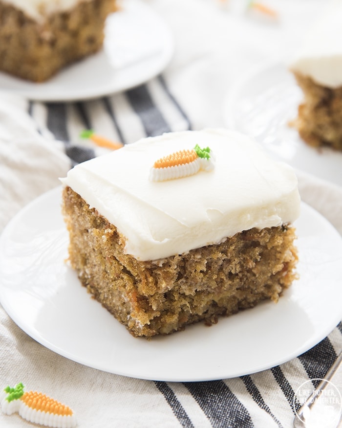 A slice of carrot cake on a small white plate. It's a square slice with a thick layer of cream cheese frosting, and topped with a candy carrot on top.