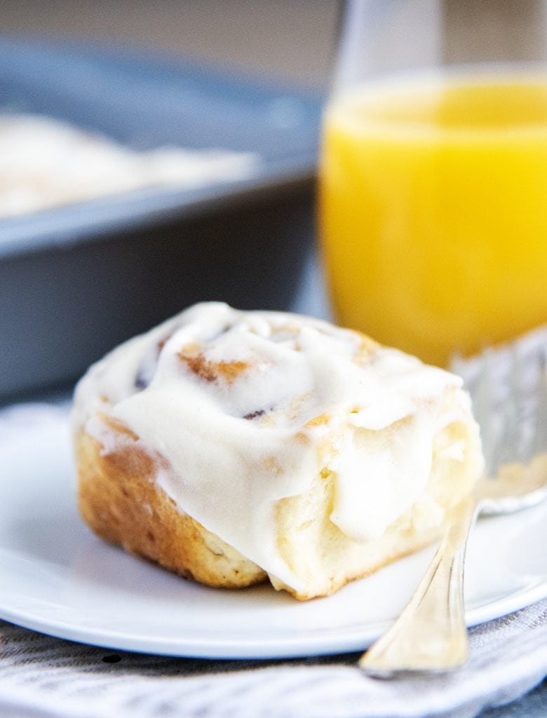 A no yeast cinnamon roll with cream cheese frosting on a plate with a glass of orange juice behind it.