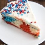 A slice of red white and blue swirled cake topped with white frosting and sprinkles.