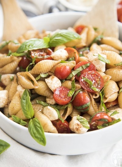 A bowl of caprese pasta salad with tomatoes and fresh basil, with two wooden spoons in the back.