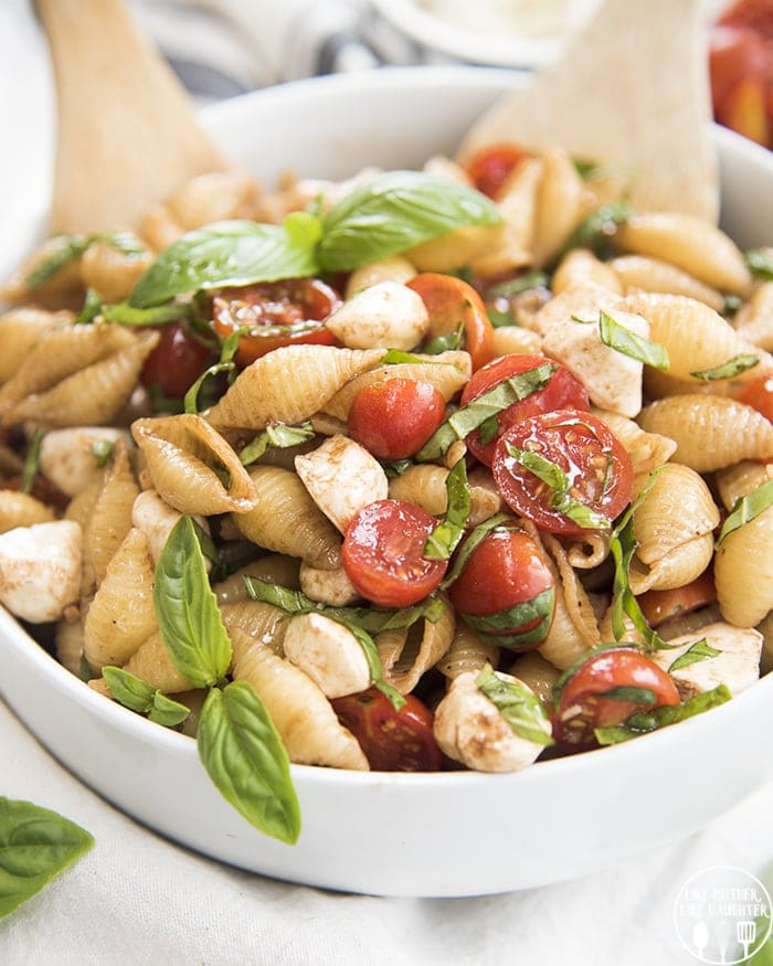 A bowl of caprese pasta salad with tomatoes and fresh basil, with two wooden spoons in the back.