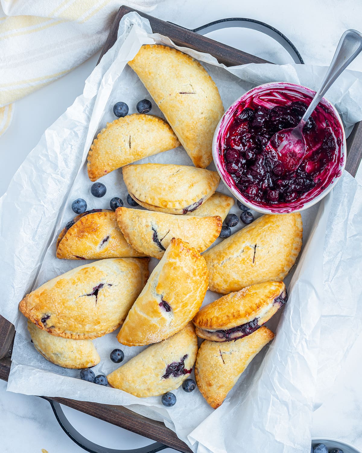 An overhead photo of a baking pan full of handheld blueberry hand pies and a bowl of blueberry sauce.
