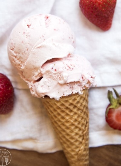 An ice cream cone with strawberry ice cream on it, and strawberries next to, all laying on a white cloth.