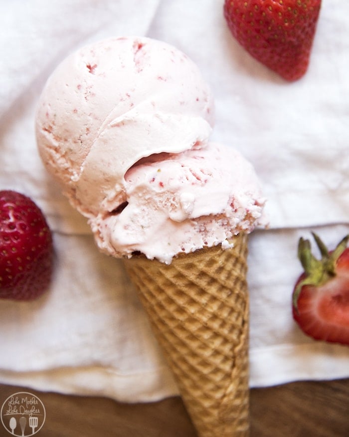 An ice cream cone with strawberry ice cream on it, and strawberries next to, all laying on a white cloth.