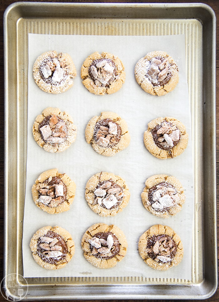 A cookie sheet topped with peanut butter cookies, with chocolate and muddy buddy pieces on top. 