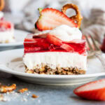A plate of strawberry pretzel salad with a layer of pretzel crust, cheesecake filling, strawberry jello, annd whipped cream on top.