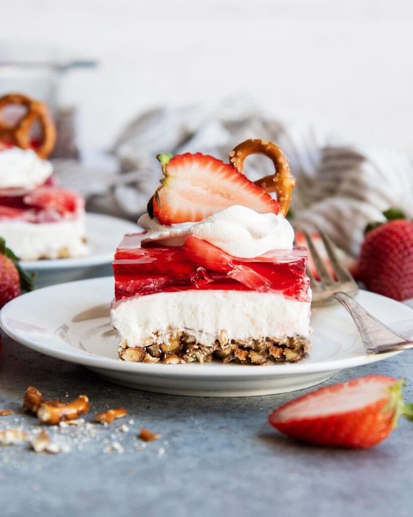 A plate of strawberry pretzel salad with a layer of pretzel crust, cheesecake filling, strawberry jello, annd whipped cream on top.