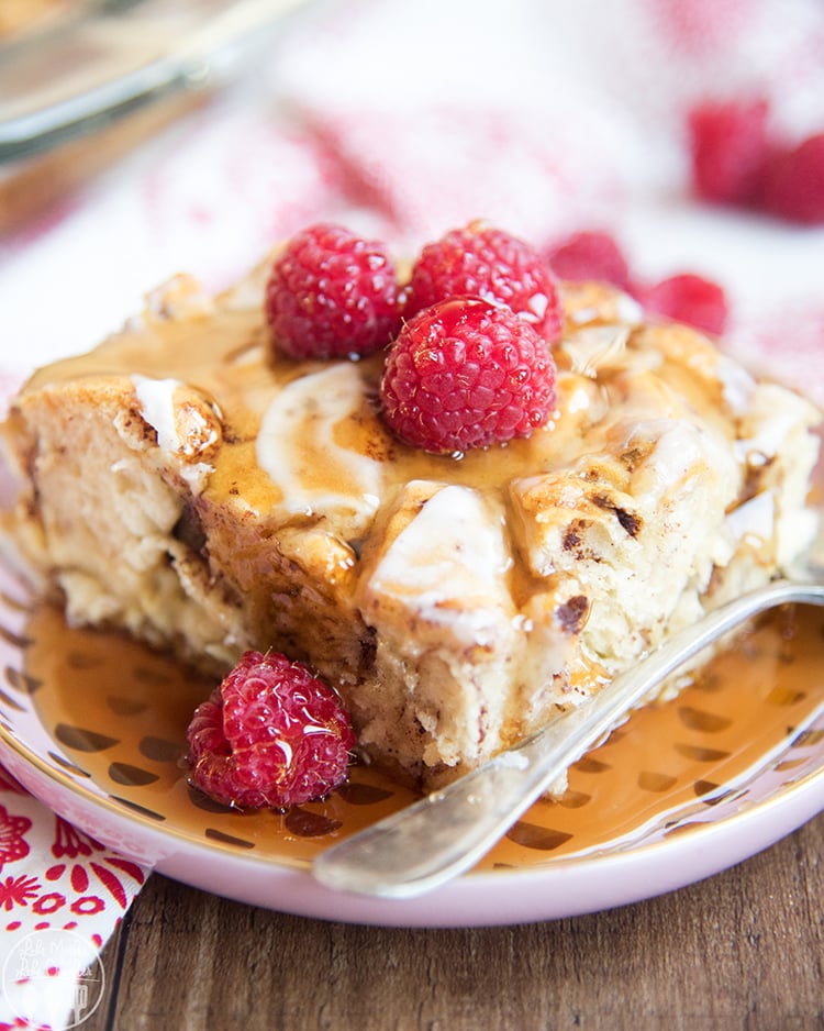 A plate of cinnamon roll french toast casserole with frosting, syrup, and raspberries on top.