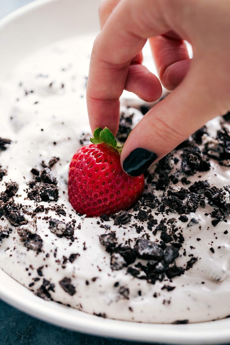 A hand dipping a strawberry into a cookies and cream fruit dip.
