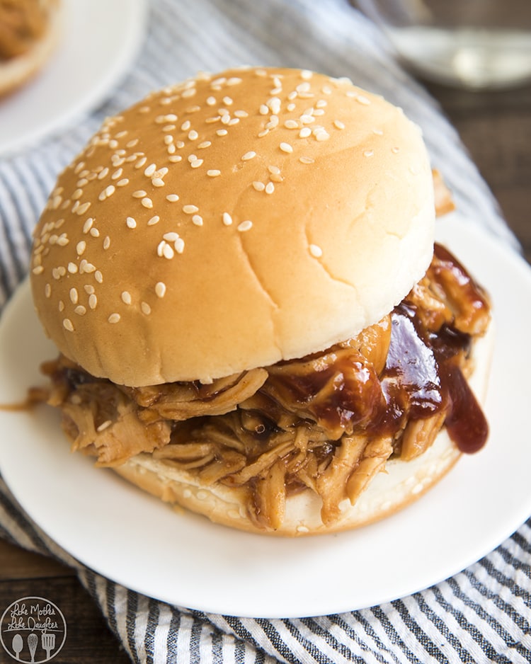 A bbq chicken sandwich on a white plate. The chicken is shredded and bbq sauce is dripping off the side, it is on a sesame bun.