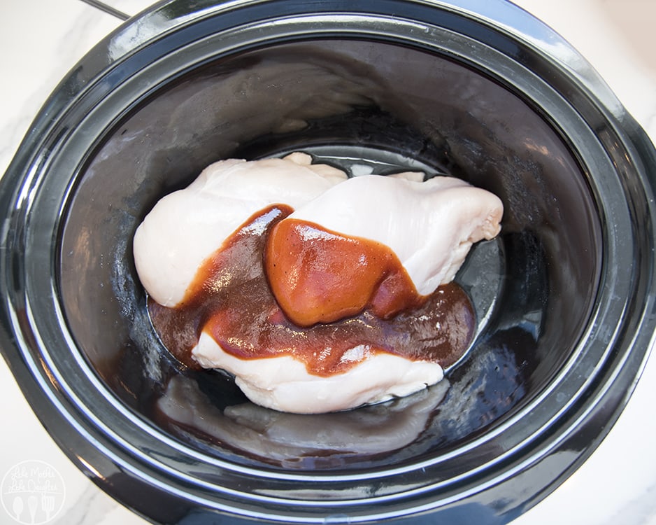 Three chicken breasts topped with barbecue sauce in a black slow cooker.