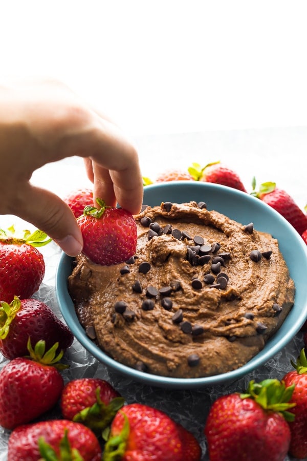 A bowl of chocolate dessert hummus with a hand dipping a strawberry into it.