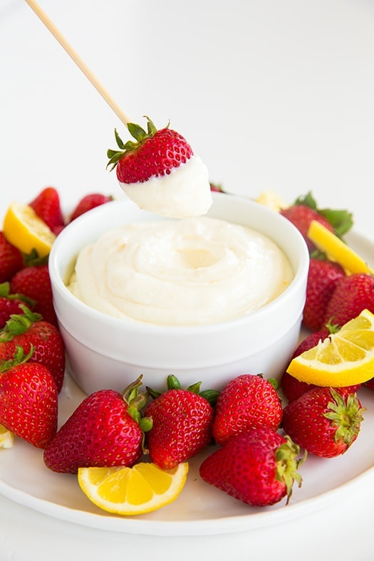 A bowl of lemon cream fruit dip with a strawberry dipping into it on a stick.