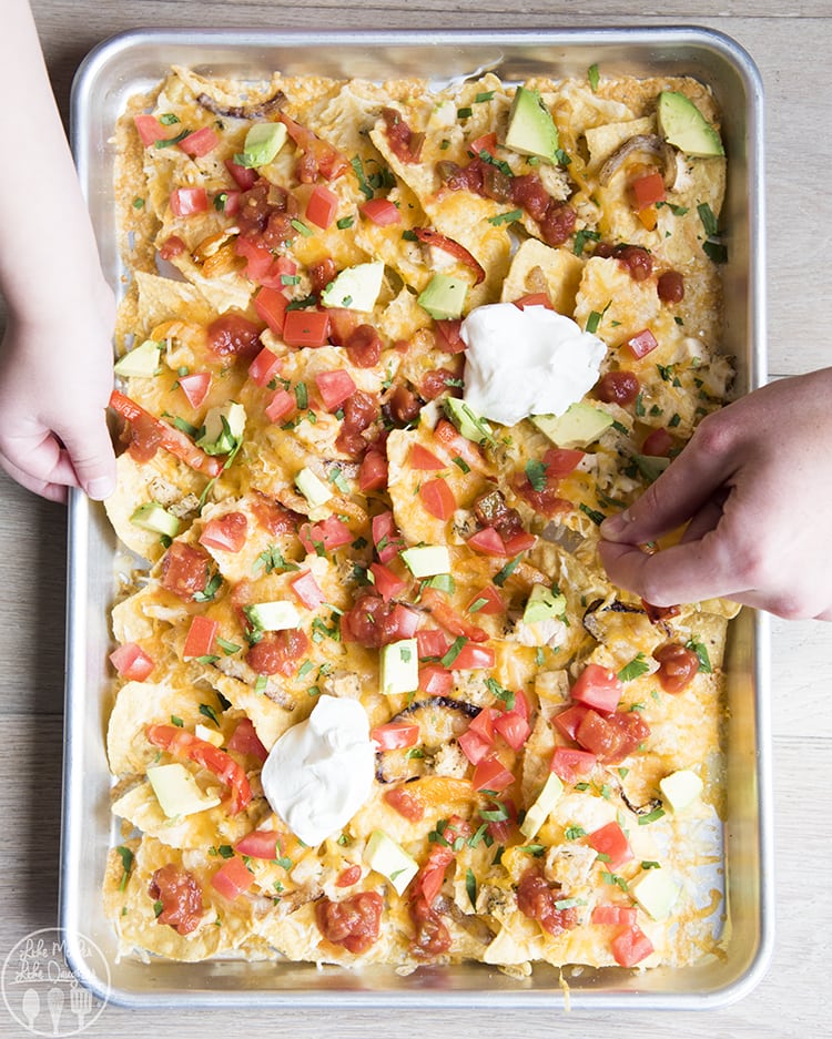 A sheet pan covered in nachos with cheese, diced tomatoes, avocado, and dollops of sour cream.