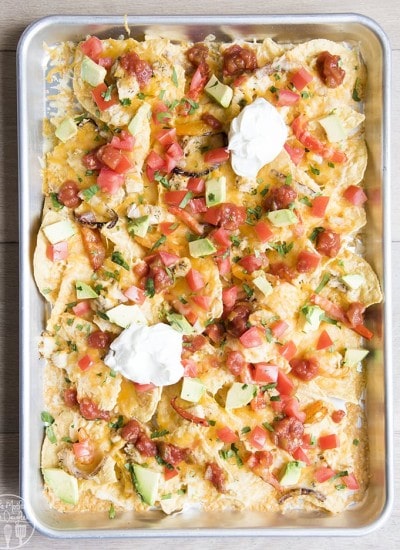 Sheet pan chicken nachos on a cookie sheet topped with tomatoes, and avocados.