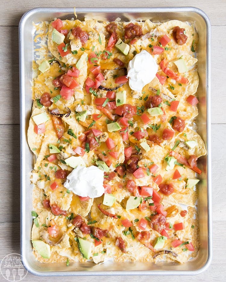 Sheet pan chicken nachos on a cookie sheet topped with tomatoes, and avocados. 