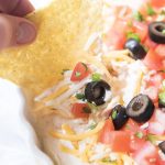 A tortilla chip dipping into a dip in a bowl covered with cheese, diced tomatoes, olives, and green onions.
