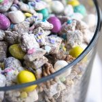A trifle dish full of Easter muddy buddies. There are pastel colored m&ms, and sprinkles, and mini marshmallows mixed in with the chocolate coated chex.