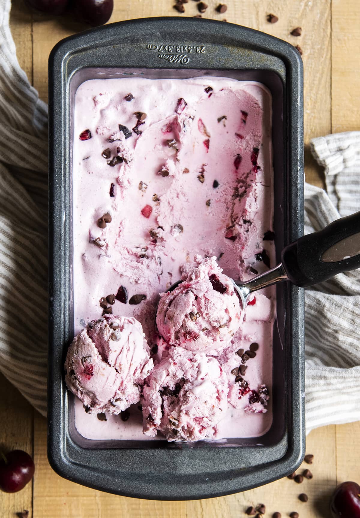 An overhead of a container of pink ice cream full of cherry pieces and chocolate chips with three scoops of it on one end.