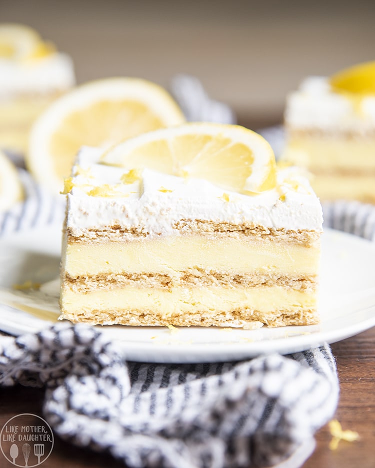 A slice of a lemon ice box cake showing layers of graham crackers and lemon pudding topped with whipped cream and a lemon wedge.