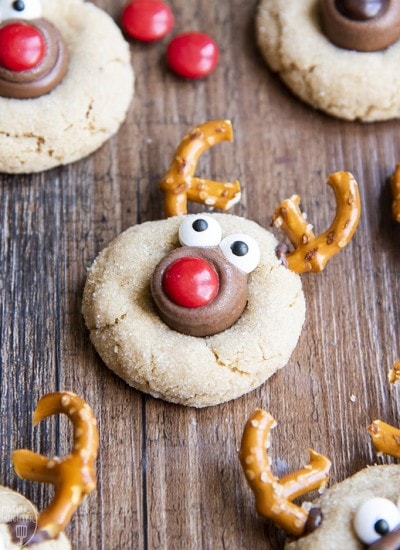 Peanut butter cookies decorated to look like a reindeer with pretzel antlers, candy eyes, and an m&m nose.