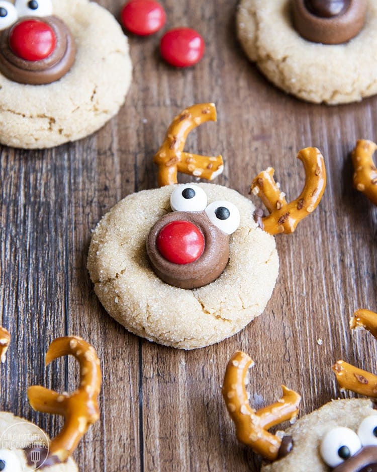 Peanut butter cookies decorated to look like a reindeer with pretzel antlers, candy eyes, and an m&m nose.