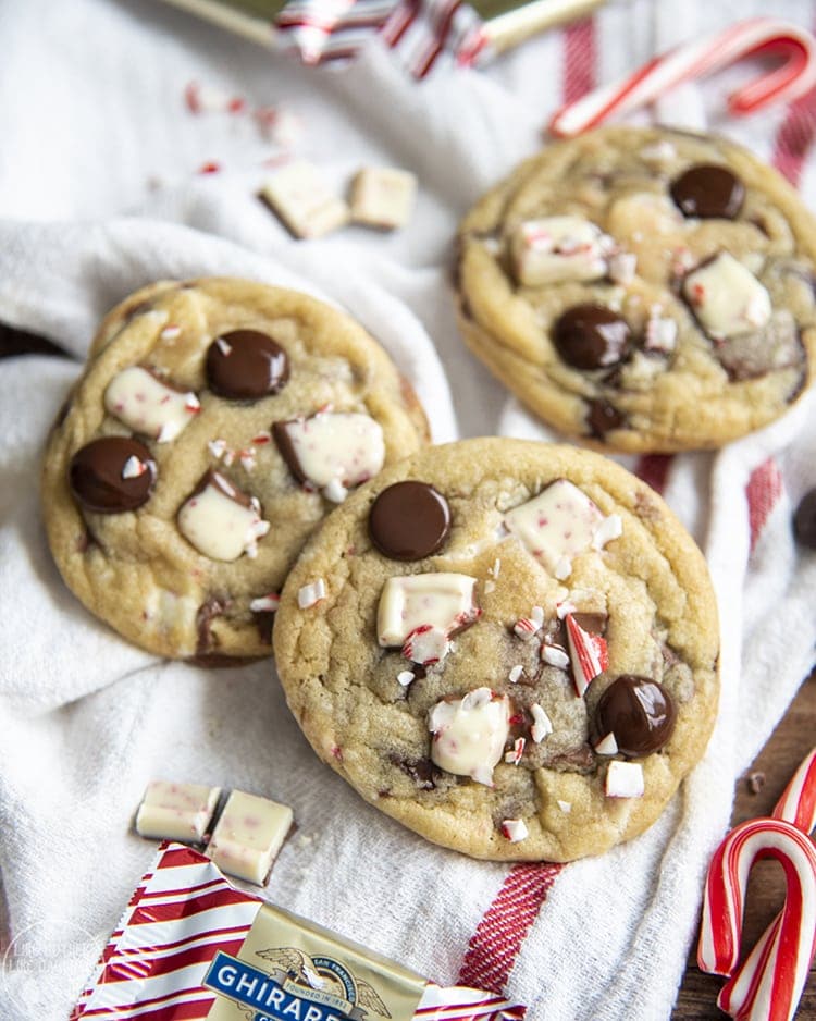 Three peppermint bark cookies on a white cloth, they are topped with pieces of chopped up peppermint bark and chocolate chips, and candy cane.