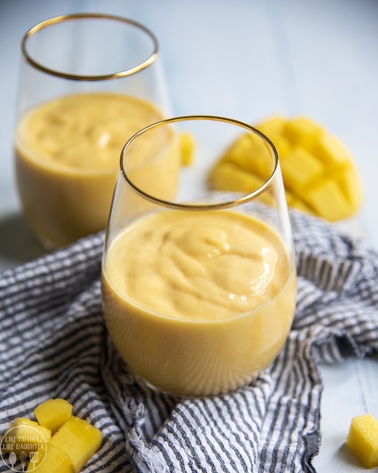 Above image of two glasses of mango smoothie with mango in the background.